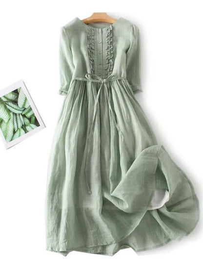 Grünes Leinenkleid mit Rüschen und Bindegürtel, langärmelig, fließend, auf Holzbügel, neben Pflanzenbild, elegantes Sommerkleid.