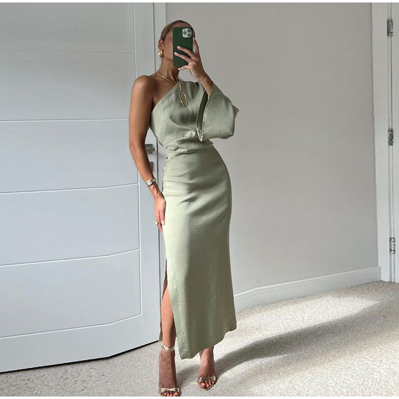 Frau in elegantem, einarmigem, olivgrünem Kleid mit hohem Schlitz posiert vor weißer Tür. Mode, Abendkleid, stilvoll, modern.
