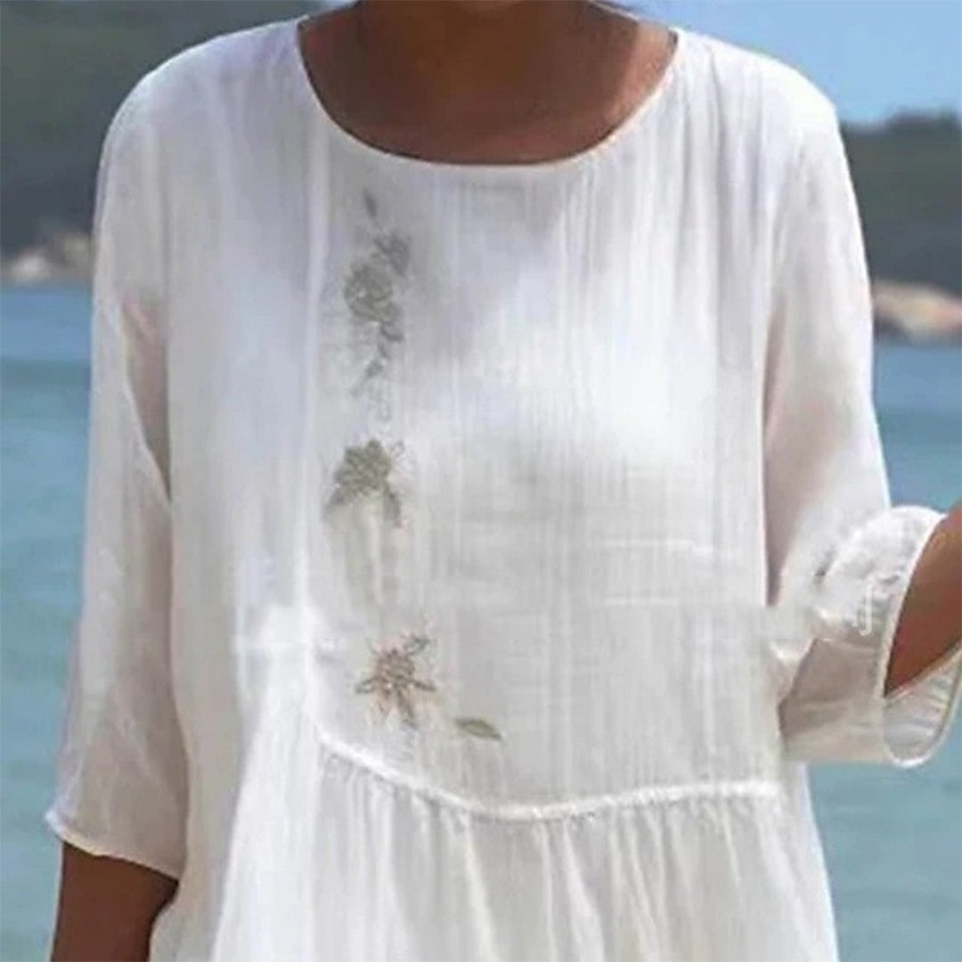 Weiße Damenbluse mit floralen Stickereien, dreiviertel Ärmel, am Strand. Sommermode, luftige Kleidung, ideal für warme Tage.