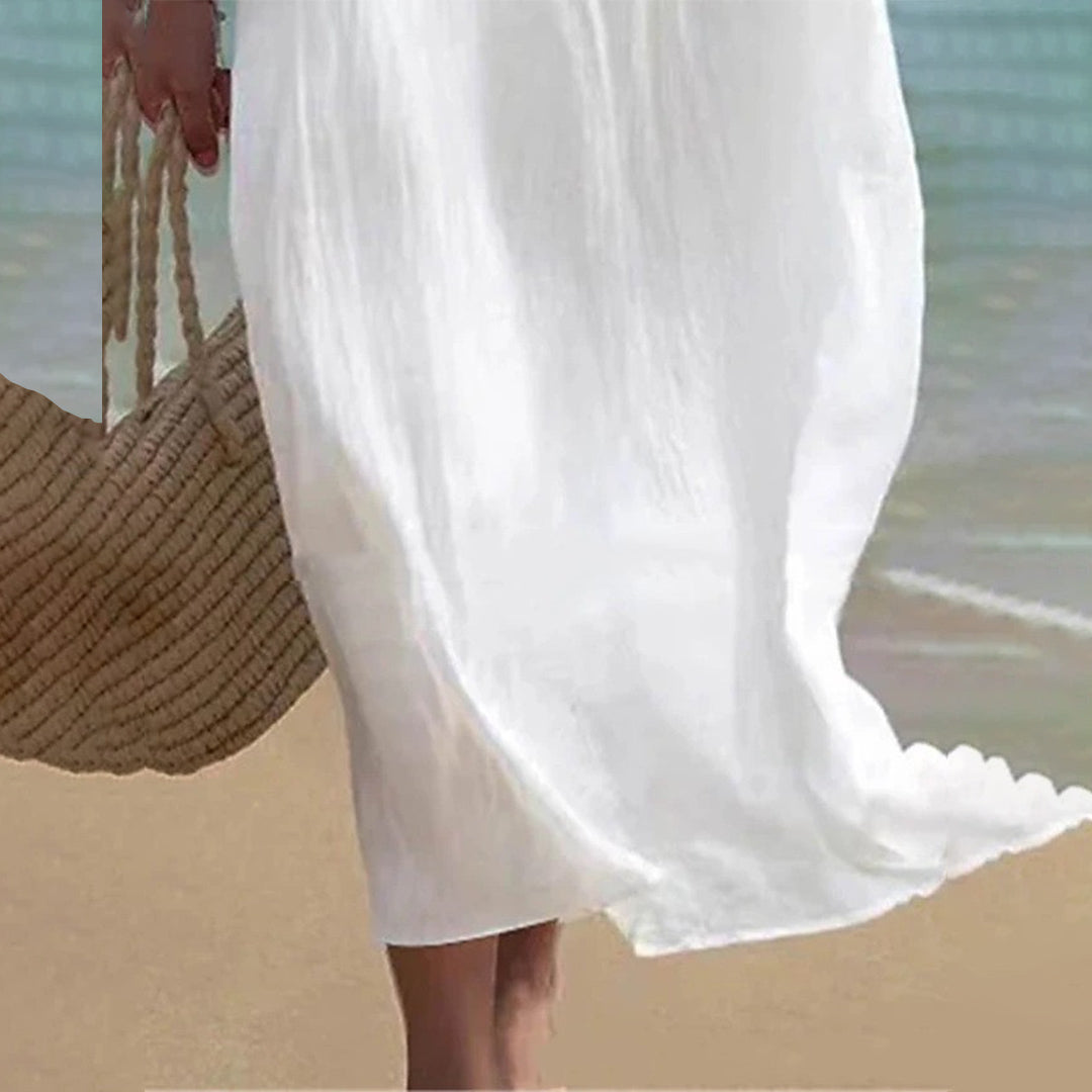 Frau in weißem Sommerkleid am Strand, hält eine Strohtasche. Leichte Brise, sandiger Hintergrund, ideal für Sommermode und Strandaccessoires.