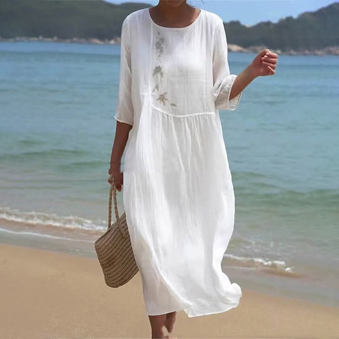 Frau in weißem Sommerkleid am Strand, trägt Strohtasche. Meer und Berge im Hintergrund. Perfekt für Sommermode, Strandkleidung, Urlaubsoutfit.
