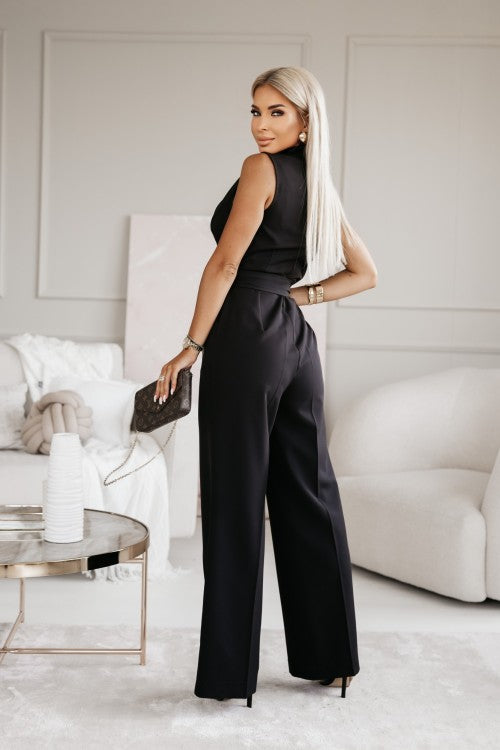 Elegante Frau in schwarzem Jumpsuit mit Handtasche, steht in modernem Wohnzimmer. Stilvolle Mode, blonde Haare, luxuriöse Inneneinrichtung.