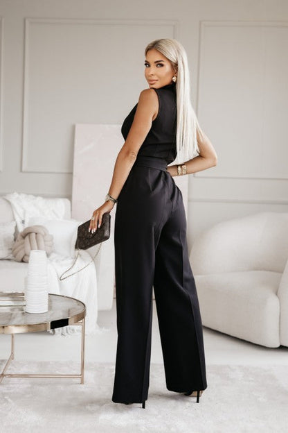 Elegante Frau in schwarzem Jumpsuit mit Handtasche, steht in modernem Wohnzimmer. Stilvolle Mode, blonde Haare, luxuriöse Inneneinrichtung.