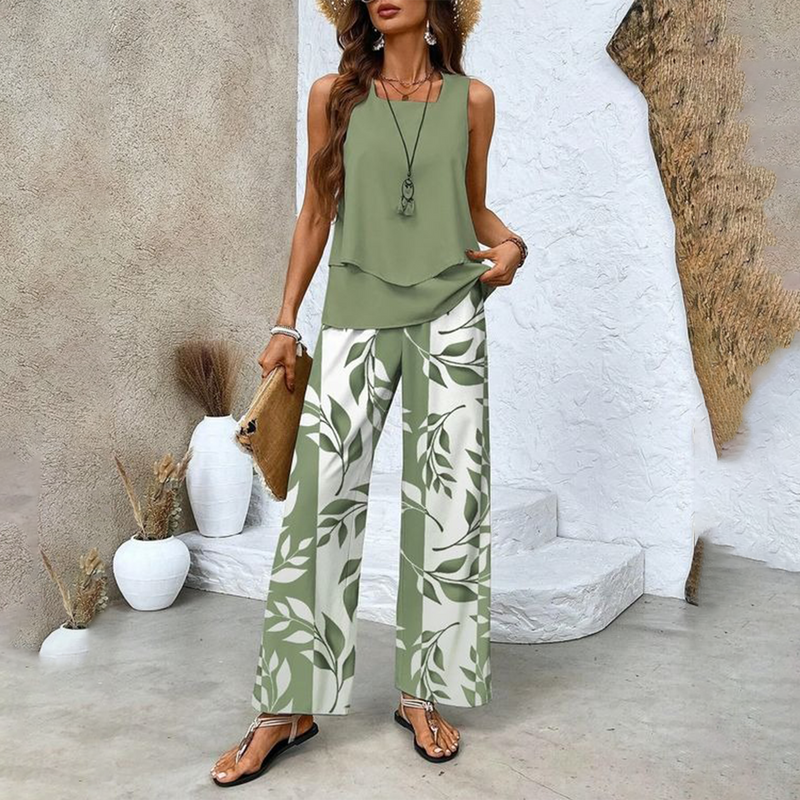 Frau in grünem ärmellosen Top und weiß-grüner Blattmusterhose, Strohhut, Sandalen, mit Clutch, vor rustikalem Hintergrund. Sommermode, Boho-Stil.