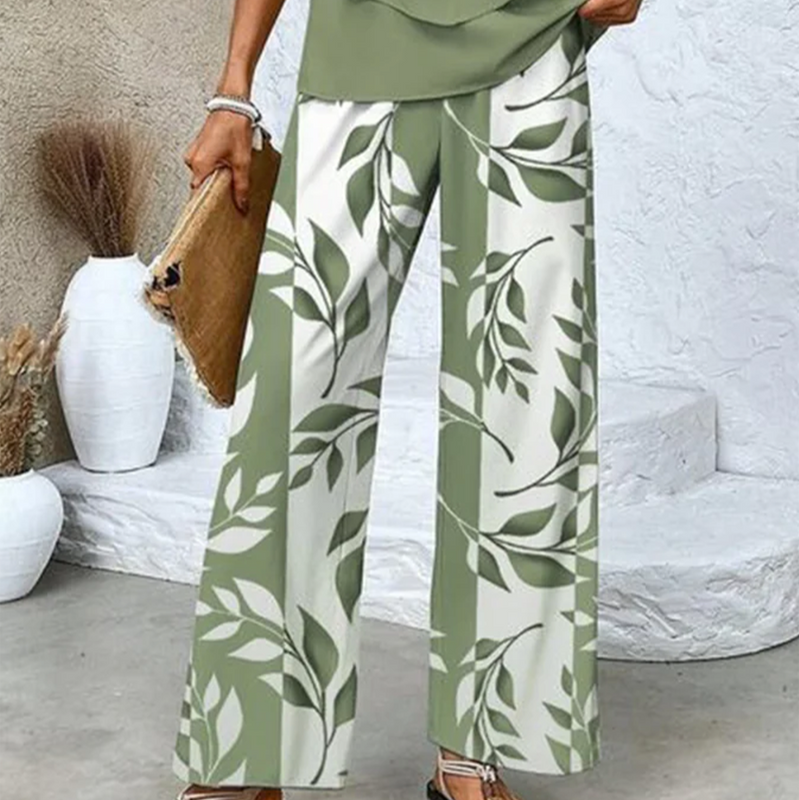 Frau in grüner, geblümter Hose, hält eine Strohtasche. Hintergrund mit weißen Vasen. Sommermode, lässiger Stil, florales Design.
