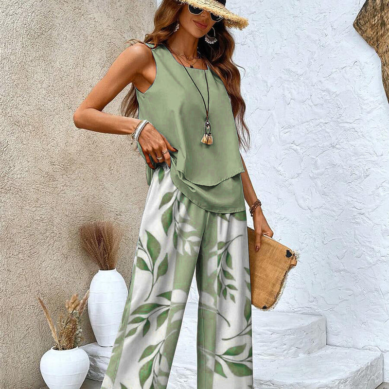 Frau in grünem ärmellosen Top und weiß-grüner Blattmusterhose, Strohhut, Sonnenbrille, mit Strohtasche, vor weißer Wand. Sommermode, Boho-Stil.