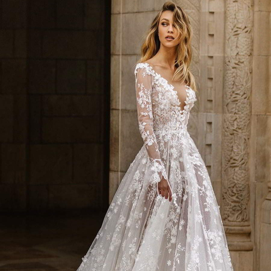 Braut in elegantem, langärmligem Spitzenkleid mit floralen Mustern vor historischer Steinwand. Hochzeitsmode, Brautkleid, romantisch, stilvoll.