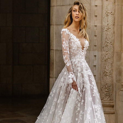 Braut in elegantem, langärmligem Spitzenkleid mit floralen Mustern vor historischer Steinwand. Hochzeitsmode, Brautkleid, romantisch, stilvoll.