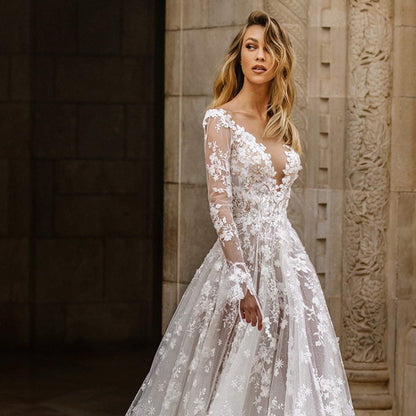 Braut in elegantem, langärmligem Spitzenkleid mit floralen Details vor historischer Steinwand. Hochzeitsmode, Brautkleid, romantisch.