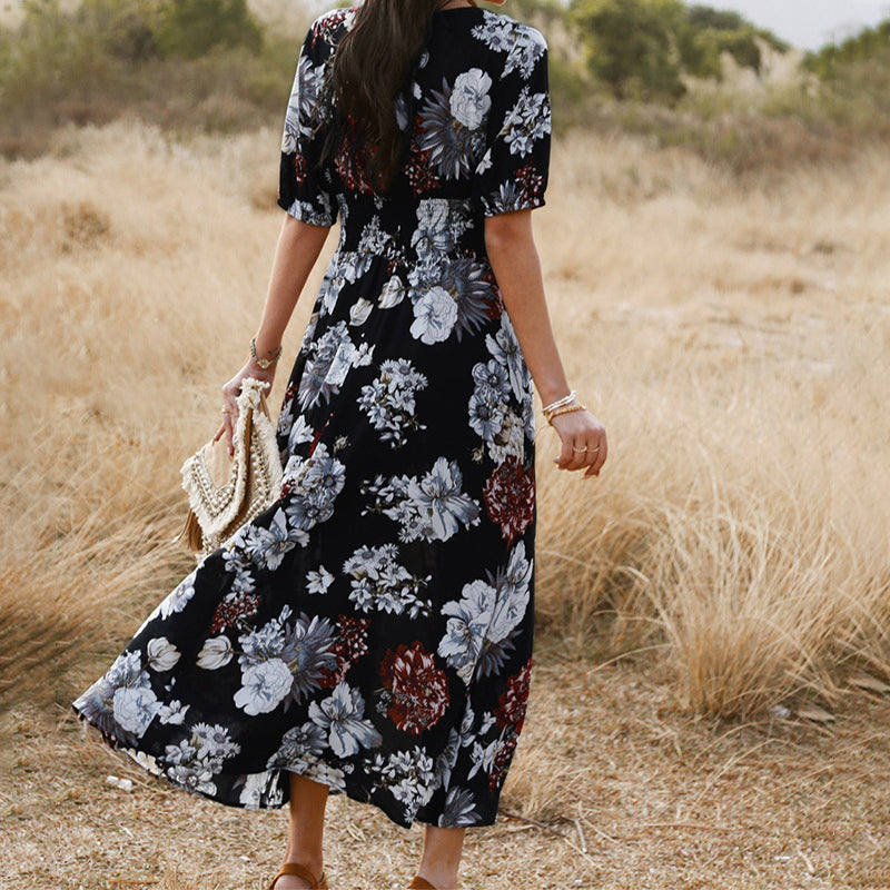 Frau in langem, geblümtem Kleid auf Feldweg, trägt Strohtasche. Sommermode, florales Design, Freizeitkleidung, Naturhintergrund.