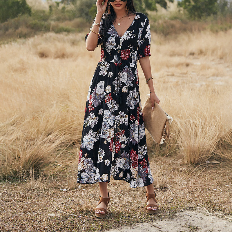 Frau in langem, geblümtem Kleid steht auf Wiese, trägt Sonnenbrille und Sandalen, hält eine Clutch. Sommermode, florales Design, Outdoor-Fotografie.