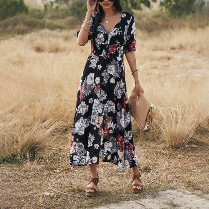 Frau in langem, geblümtem Kleid steht auf Wiese, trägt Sonnenbrille und Sandalen, hält eine Clutch. Sommermode, florales Design, Outdoor-Fotografie.