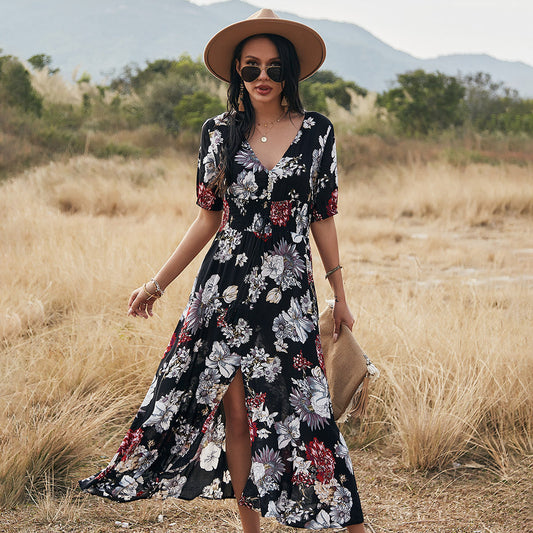 Frau in floralem Maxikleid und Hut steht auf Wiese vor Bergkulisse. Sommermode, Boho-Stil, Outdoor-Fotografie.