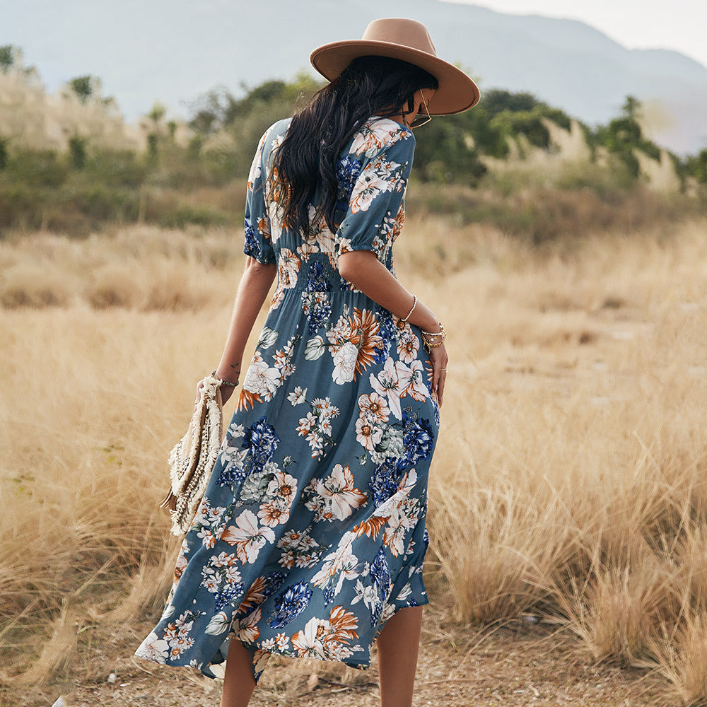 Frau im blauen, geblümten Kleid und Hut spaziert durch eine trockene Wiesenlandschaft. Sommermode, Boho-Stil, Natur, Freizeitkleidung.