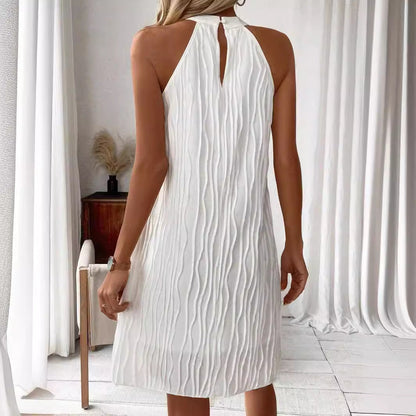 Frau in elegantem, weißem, ärmellosem Kleid mit strukturiertem Muster, steht in modernem, minimalistischem Raum mit Vorhängen und Holzmöbeln.