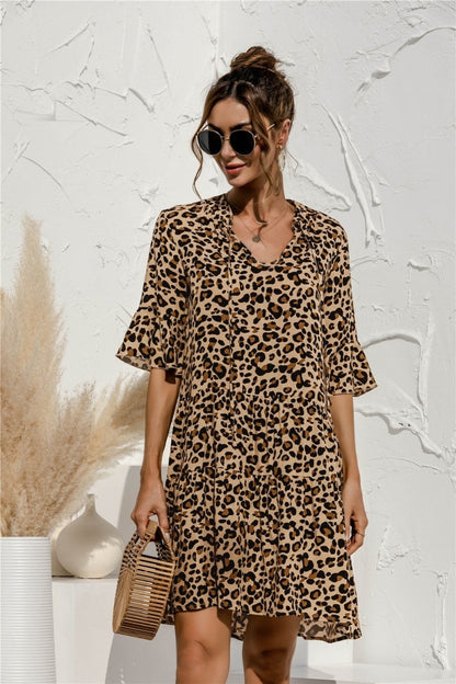 Frau in einem Leopardenmuster-Kleid mit Sonnenbrille, hält eine Bambustasche, vor weißer Wand mit Pampasgras. Modetrend, Sommerkleid, Damenmode.