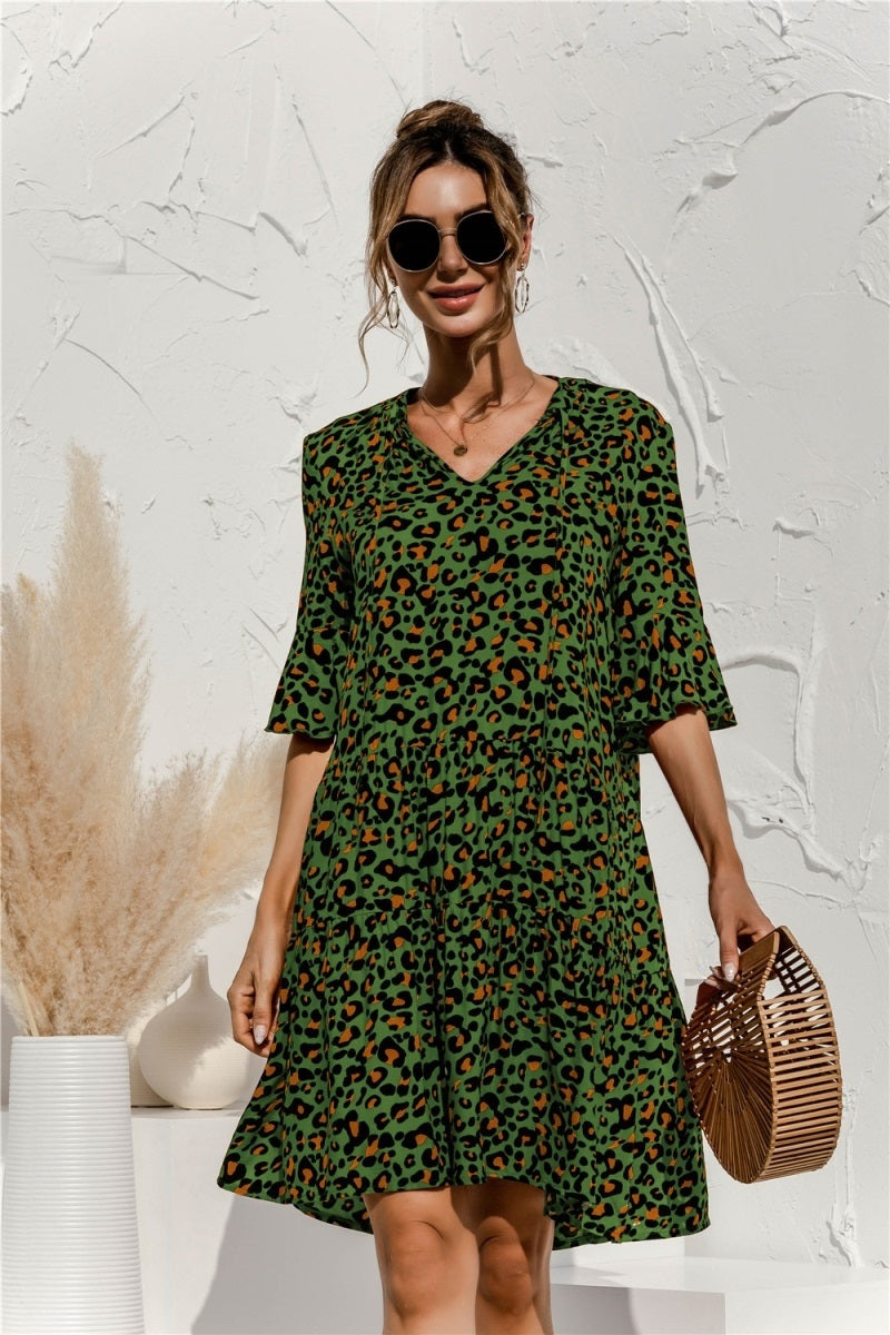 Frau in grünem Leopardenkleid mit Sonnenbrille, hält Bambustasche. Hintergrund mit Pampasgras und weißer Wand. Modetrend, Sommerkleid, Damenmode.
