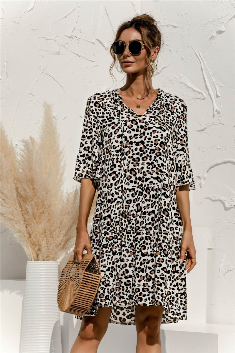 Frau in einem modischen Leopardenmuster-Kleid mit Sonnenbrille, hält eine Bambustasche. Eleganter Sommerlook vor weißer Wand mit Pampasgras.