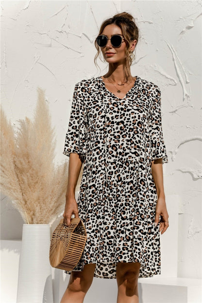 Frau in einem modischen Leopardenmuster-Kleid mit Sonnenbrille, hält eine Bambustasche. Eleganter Sommerlook vor weißer Wand mit Pampasgras.