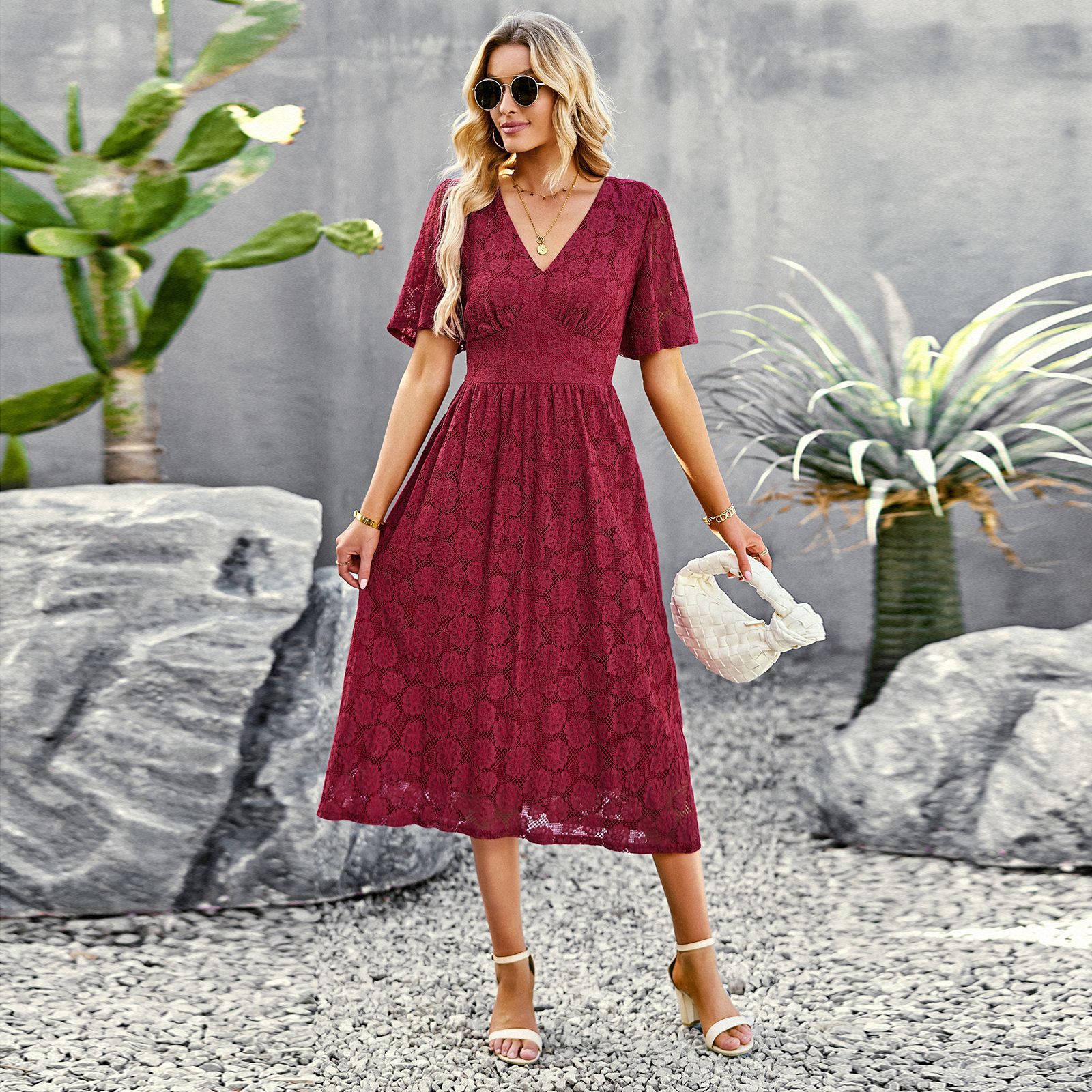 Frau in rotem Spitzenkleid, Sonnenbrille und Sandalen posiert im Freien neben Kakteen und Felsen. Sommermode, elegantes Outfit, Freizeitkleidung.