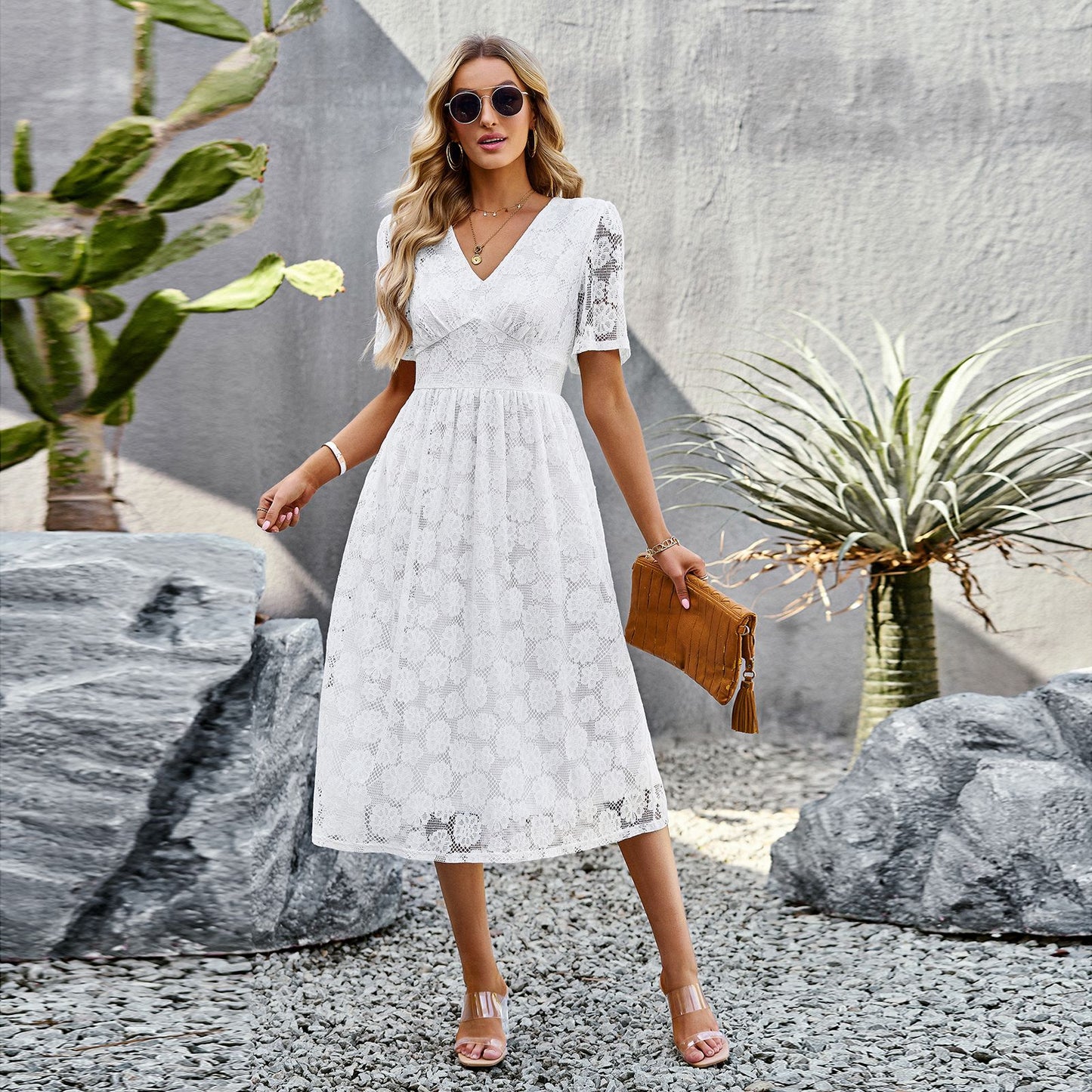 Frau in weißem Spitzenkleid, Sonnenbrille und Sandalen, steht zwischen Kakteen und Felsen, hält eine braune Clutch. Sommermode, elegantes Outfit.