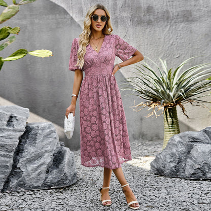 Frau in rosa Spitzenkleid, Sonnenbrille und Sandalen, steht zwischen Steinen und Pflanzen. Sommermode, elegantes Outfit, Outdoor-Fotoshooting.