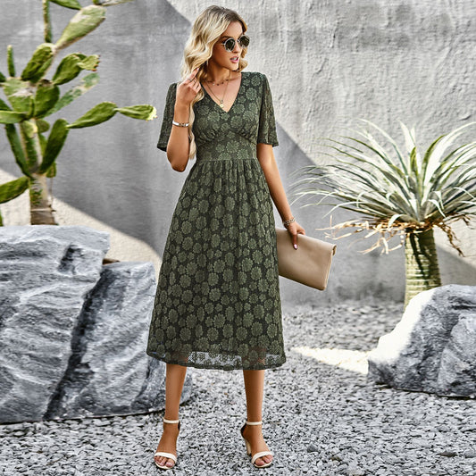 Elegante Frau im grünen Spitzenkleid, Sonnenbrille, mit Clutch, steht auf Kiesweg vor Kakteen und Agave. Sommermode, Damenkleid, Outdoor-Fotoshooting.