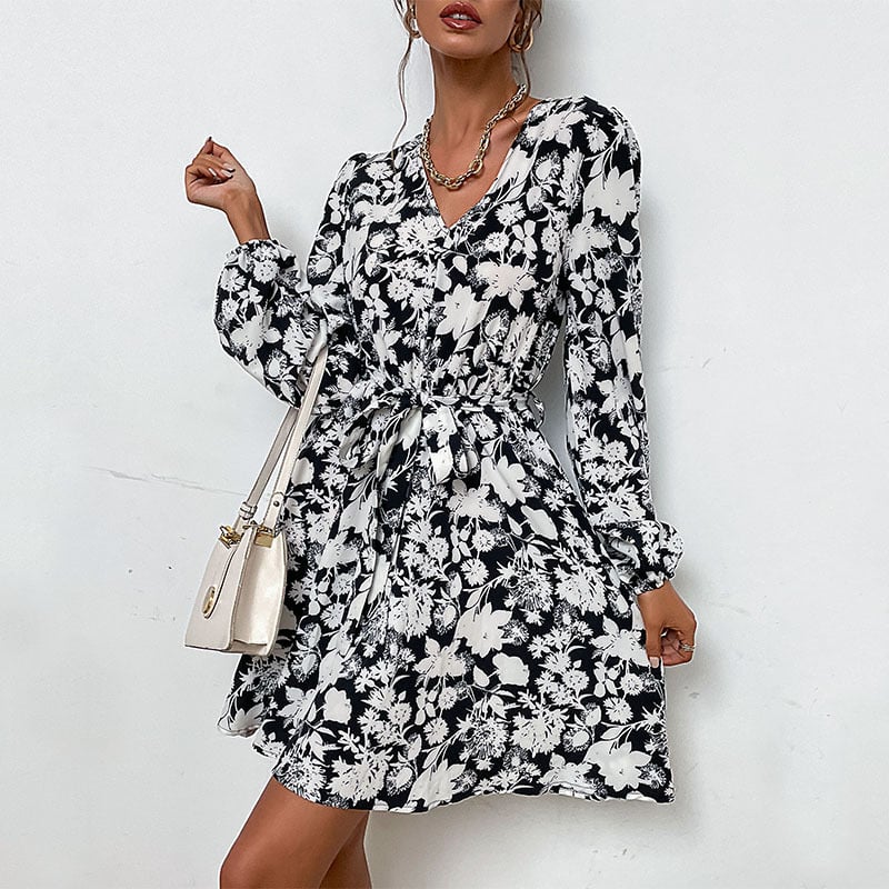 Schwarz-weißes geblümtes Kleid mit langen Ärmeln und V-Ausschnitt, kombiniert mit einer cremefarbenen Handtasche. Elegante Damenmode, stilvoll und modern.