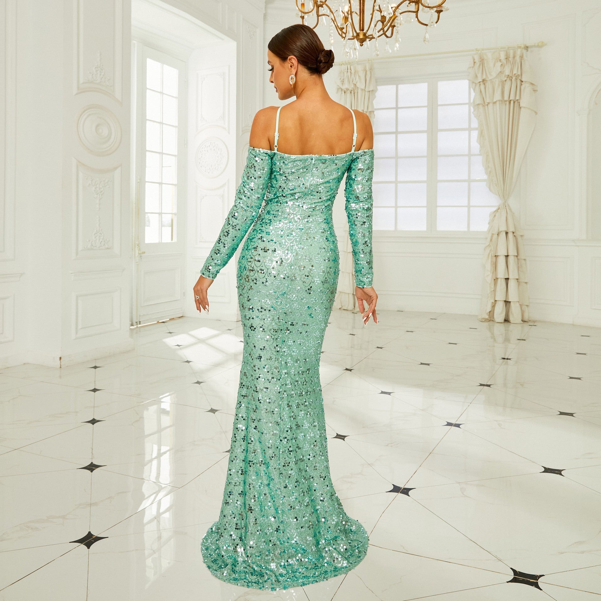 Frau in eleganter, schulterfreier, grüner Paillettenkleid, steht in luxuriösem, hellen Raum mit Kronleuchter und Marmorboden, Rückansicht.