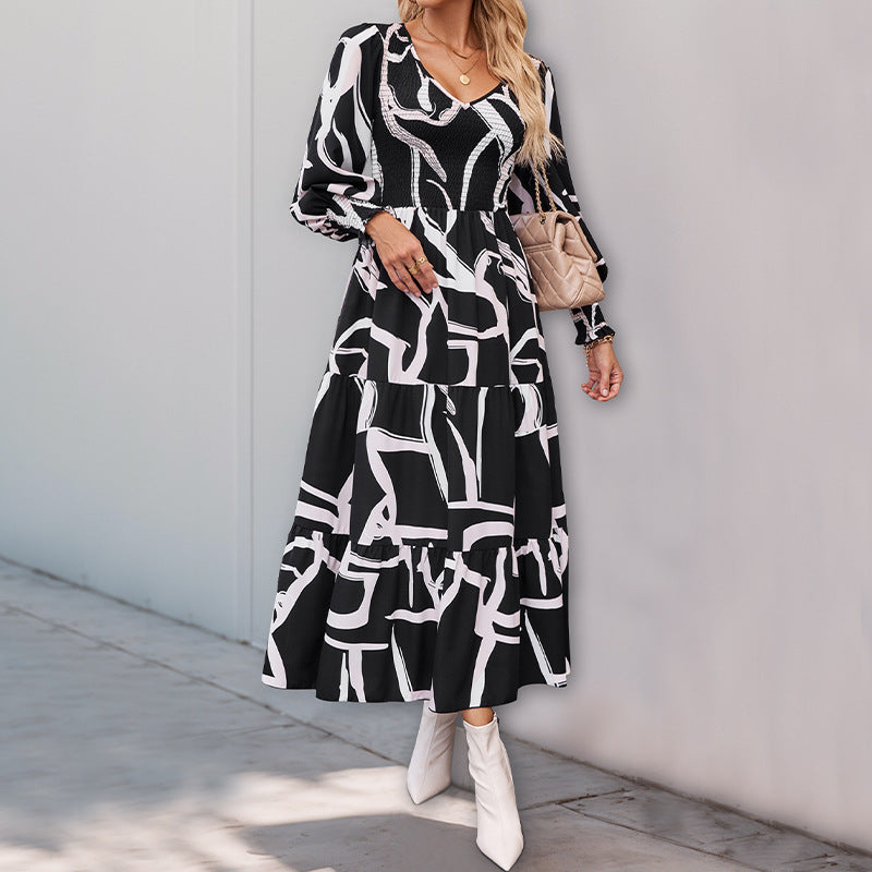Frau in schwarz-weiß gemustertem Maxikleid, trägt weiße Stiefel und beige Handtasche, steht vor grauer Wand. Modetrend, eleganter Look.