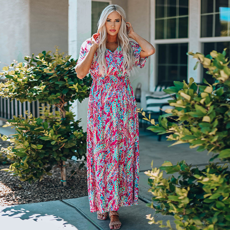 Frau in buntem Maxikleid steht im Garten, umgeben von grünen Büschen. Sommermode, florales Design, Freizeitkleidung, stilvolle Frau.