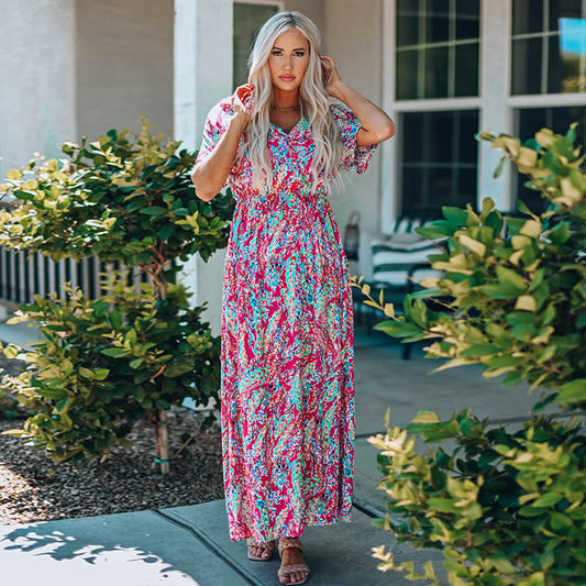 Frau in buntem Maxikleid steht im Garten, umgeben von grünen Büschen. Sommermode, florales Design, Freizeitkleidung, stilvolle Frau.