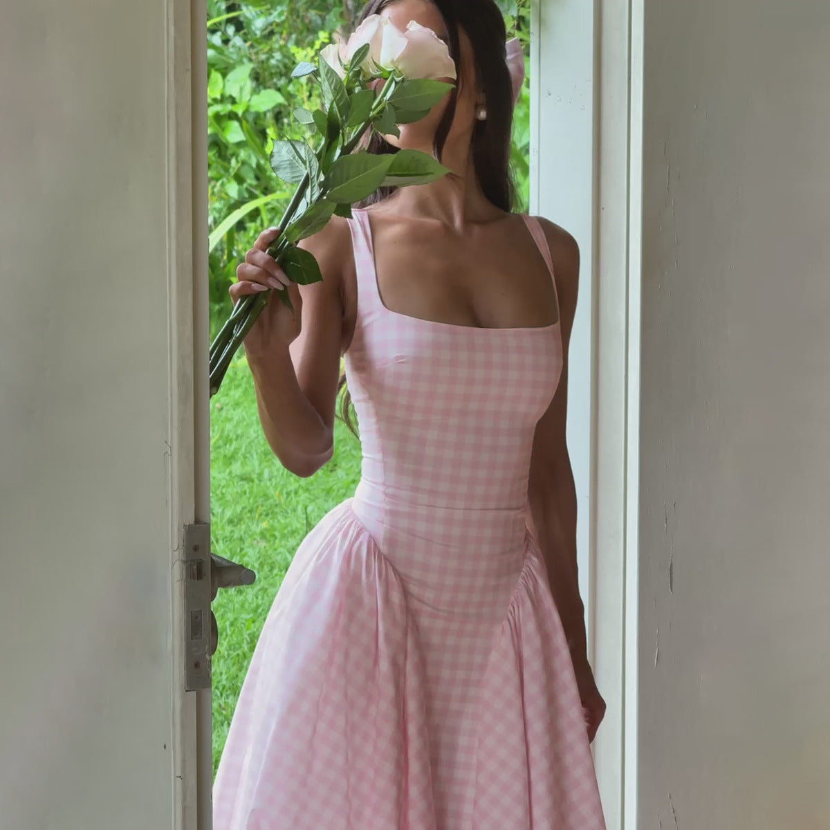 Frau in rosa kariertem Kleid hält Blume, steht in Türrahmen, grüner Garten im Hintergrund. Sommermode, elegantes Kleid, Blumenstrauß.