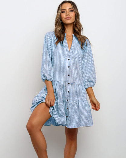 Frau in blau-weiß kariertem Kleid mit Knopfleiste, Dreiviertelärmeln und Rüschen, lächelt vor neutralem Hintergrund. Modetrend, Sommerkleid.
