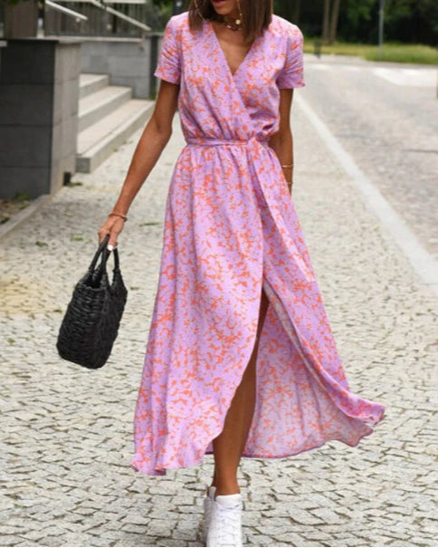 Frau in einem lila-blumigen Wickelkleid mit schwarzen Korbtasche und weißen Sneakers auf gepflastertem Weg. Sommermode, lässiger Stil.