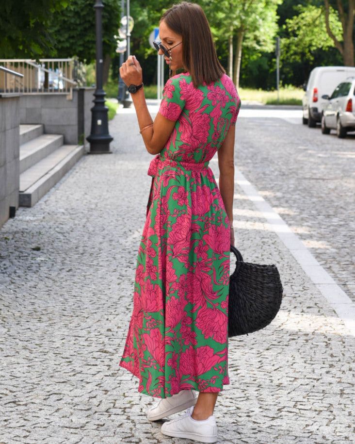 Frau in langem, geblümtem Kleid und weißen Sneakers auf gepflasterter Straße, hält schwarzen Korb. Sommermode, Streetstyle, urbane Eleganz.