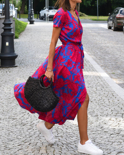 Frau in rotem und blauem Blumenkleid mit schwarzer Tasche und weißen Sneakers auf gepflastertem Gehweg. Sommermode, Straßenstil, Damenkleidung.
