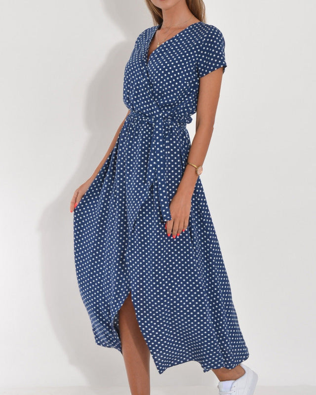 Frau in blauem, gepunktetem Sommerkleid, knielang, mit kurzen Ärmeln und V-Ausschnitt, trägt weiße Sneaker. Elegantes Freizeitoutfit.