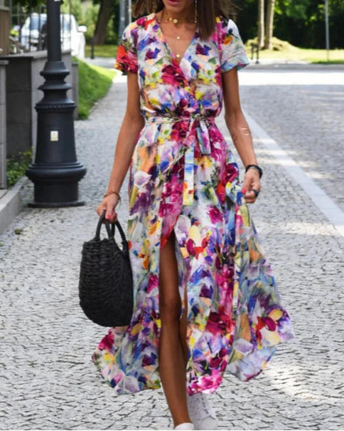 Frau in buntem, geblümtem Maxikleid mit Gürtel, trägt schwarze Korbtasche, spaziert auf gepflastertem Weg. Sommermode, Freizeitkleidung, Straßenstil.