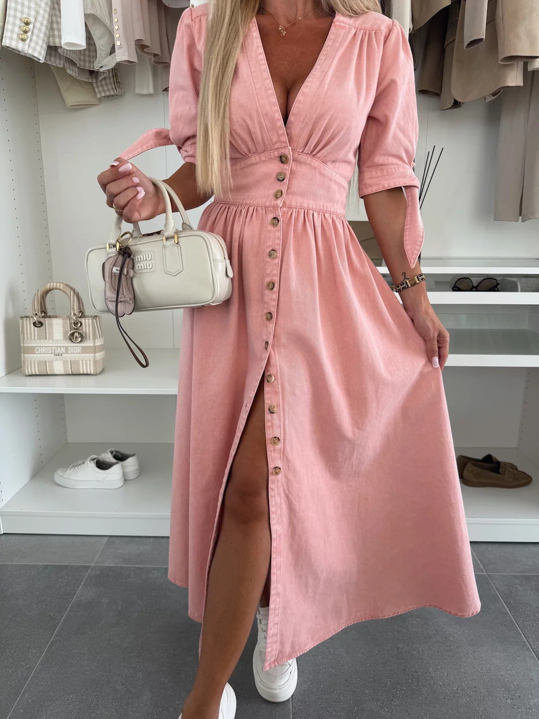 Frau in rosa Knopfkleid mit weißer Handtasche, vor einem Kleiderschrank mit beigen und weißen Kleidungsstücken. Elegante Mode, Sommerkleid.