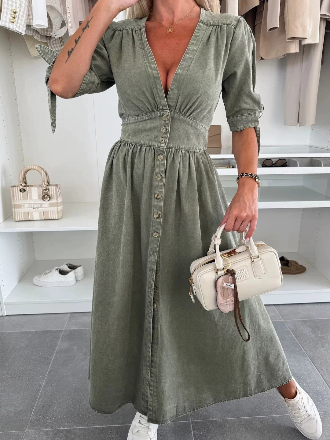 Frau in olivgrünem Kleid mit Knopfleiste, hält cremefarbene Handtasche, trägt weiße Sneaker. Mode, Damenbekleidung, eleganter Look.