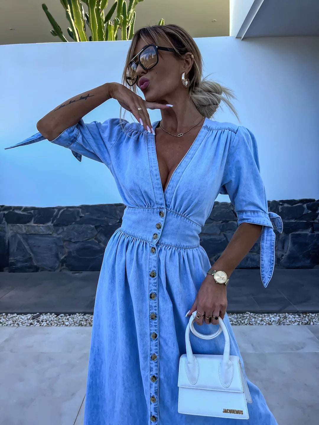 Frau in blauem Jeanskleid mit Knopfleiste, hält weiße Jacquemus-Tasche, trägt Sonnenbrille und goldene Accessoires, vor modernem Hintergrund.