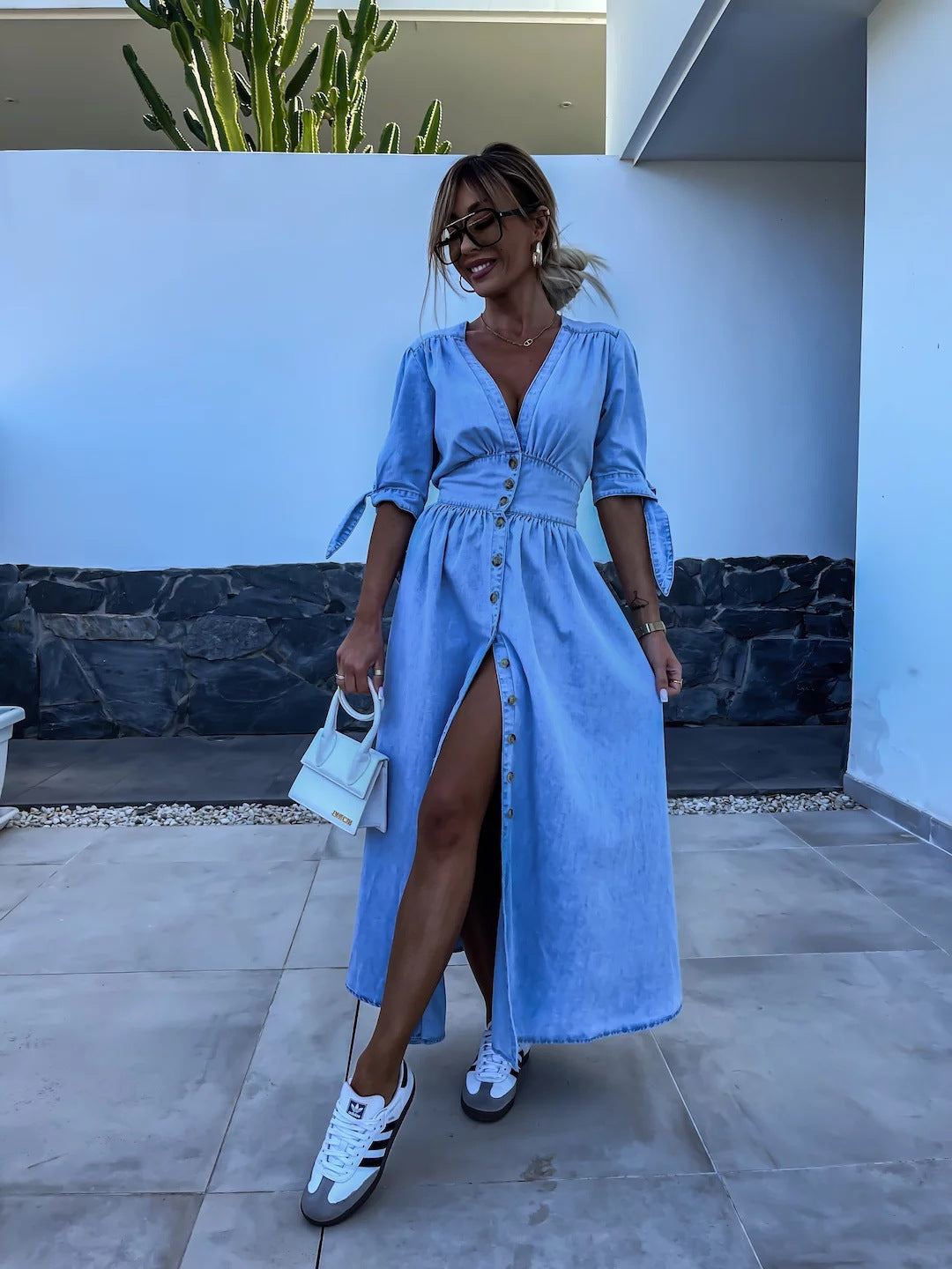 Frau in blauem Jeanskleid mit Knopfleiste und weißer Handtasche, trägt weiße Sneaker, steht vor modernem Haus mit Kaktus im Hintergrund.