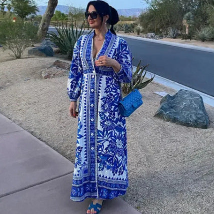 Frau in blau-weißem Maxikleid mit Blumenmuster, steht auf Gehweg in Wüstenlandschaft, trägt blaue Sandalen und blaue Tasche.