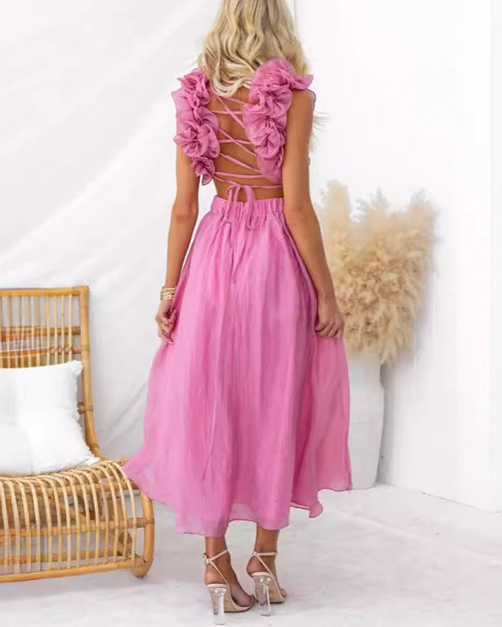 Blonde Frau in rosa Sommerkleid mit Rückenausschnitt und Rüschen, steht neben einem Rattan-Stuhl. Elegante Mode, femininer Stil, Sommerkleid.