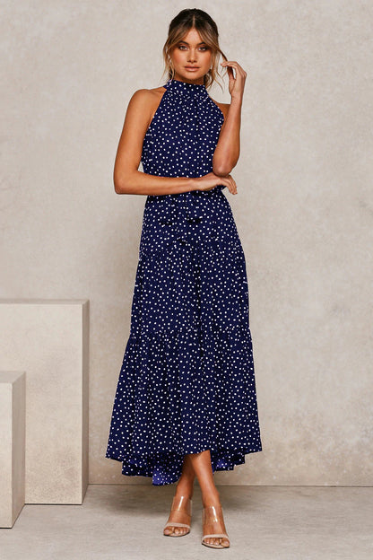 Frau in dunkelblauem, ärmellosem Maxikleid mit weißen Punkten, stehend vor neutralem Hintergrund. Elegante Mode, Sommerkleid, Polka-Dot-Trend.