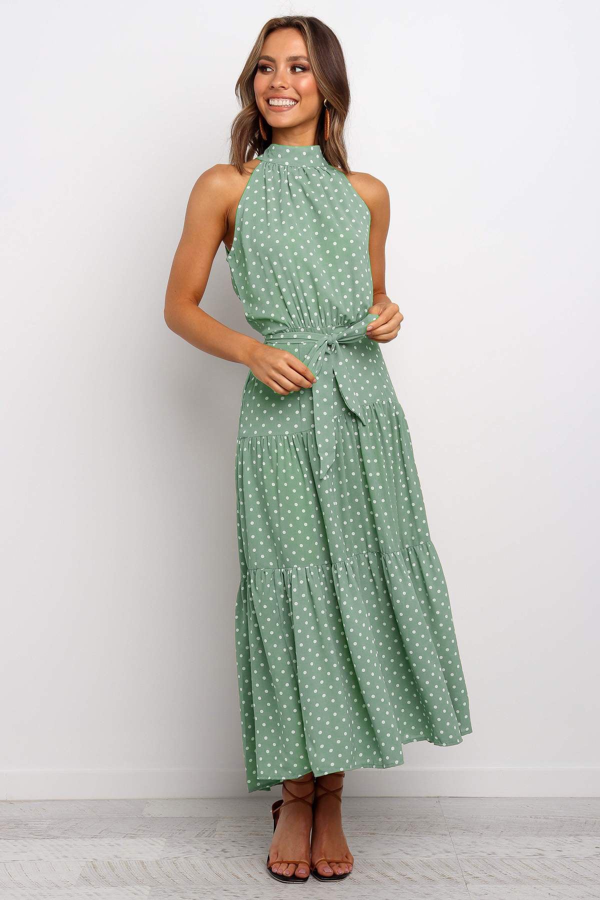 Frau in grünem, ärmellosem Maxikleid mit weißen Punkten, lächelt und steht vor weißem Hintergrund. Sommermode, elegantes Damenoutfit, gepunktetes Kleid.