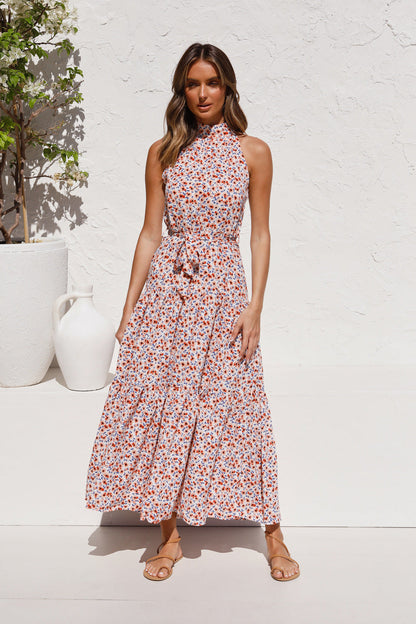 Frau in geblümtem Sommerkleid vor weißer Wand, neben großer Vase. Elegantes, ärmelloses Maxikleid, ideal für Sommermode und Freizeit.