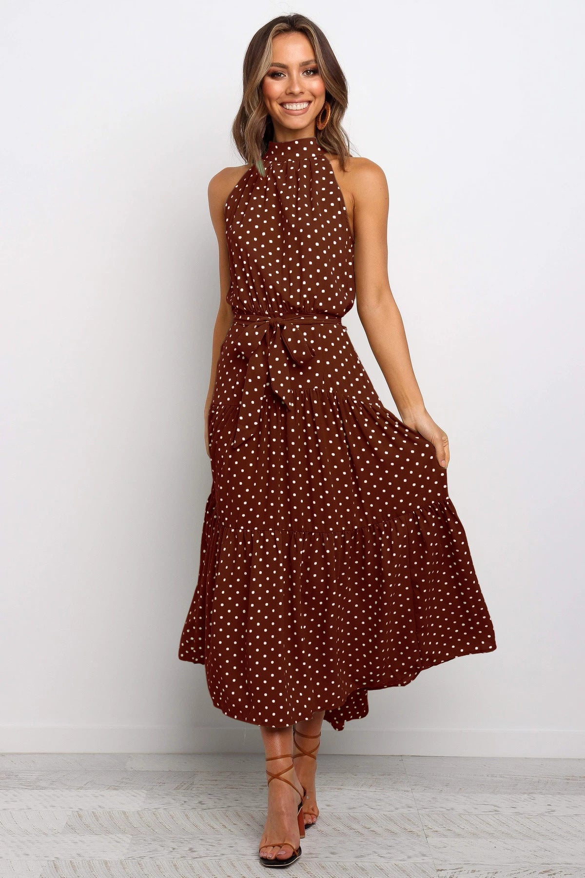 Frau in braunem, ärmellosem Kleid mit weißen Punkten, lächelt und steht auf hellem Hintergrund. Elegantes Sommeroutfit, modisches Design.