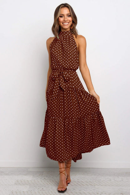 Frau in braunem, ärmellosem Kleid mit weißen Punkten, lächelt und steht auf hellem Hintergrund. Elegantes Sommeroutfit, modisches Design.
